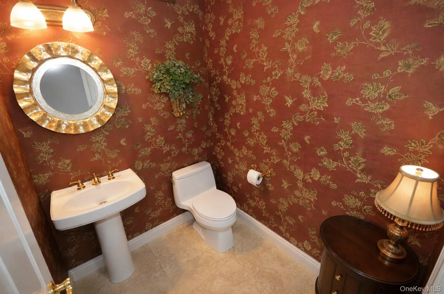 Pedestal sink with brass-toned fixtures and an ornate round mirror Pedestal sink with brass-toned fixtures and an ornate round mirror