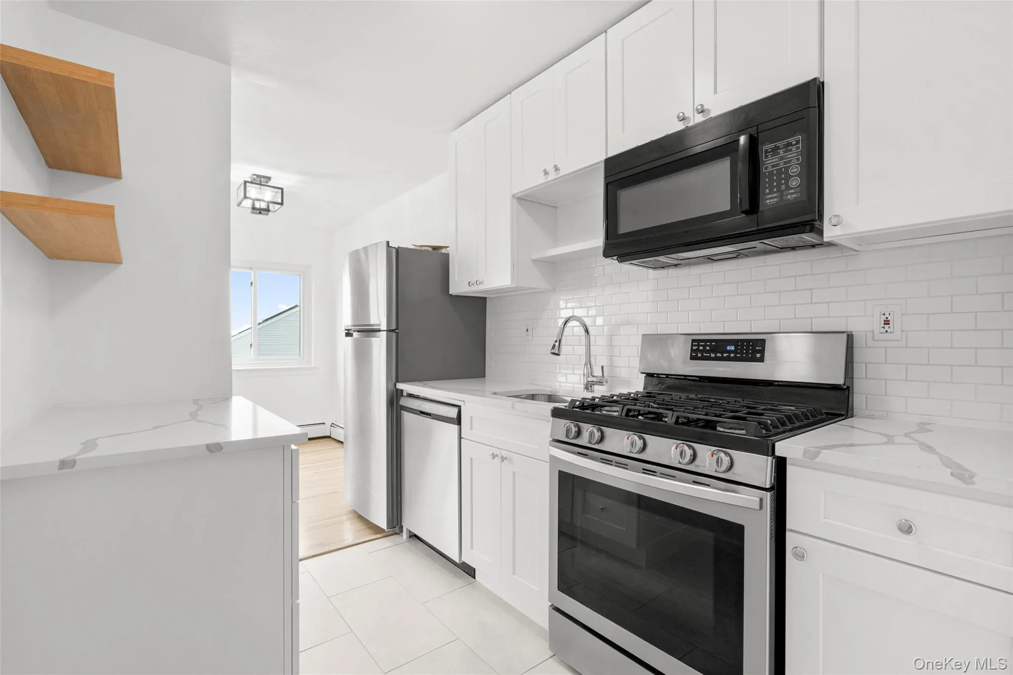 View of kitchen from entry way. Kitchen features stainless steel appliances, tile floor and quartz countertops and floating wood shelves. Gas stove, dishwasher, refrigerator and microwave. View of kitchen from entry way. Kitchen features stainless steel appliances, tile floor and quartz countertops and floating wood shelves. Gas stove, dishwasher, refrigerator and microwave.