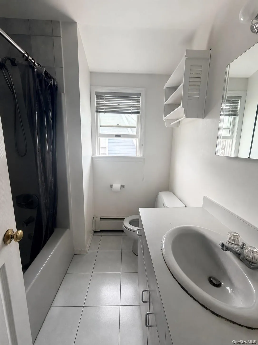 Bathroom featuring a white ceramic tile floor, a shower-tub combination with tiled walls, a window with blinds, a wall-mounted shelving unit, and a vanity with an integrated sink Bathroom featuring a white ceramic tile floor, a shower-tub combination with tiled walls, a window with blinds, a wall-mounted shelving unit, and a vanity with an integrated sink
