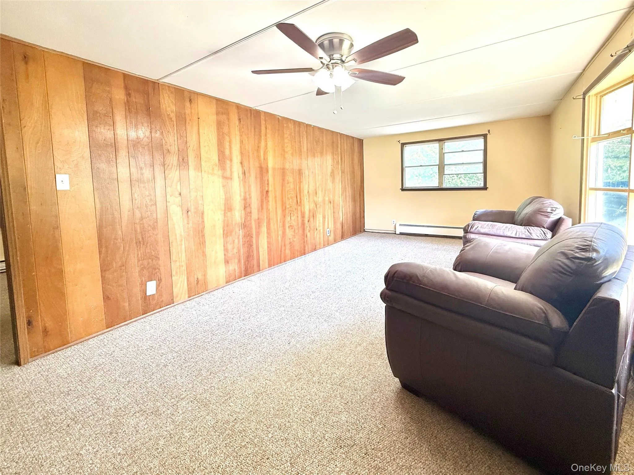 Carpeted living room featuring baseboard heating, wooden walls, healthy amount of natural light, and ceiling fan Carpeted living room featuring baseboard heating, wooden walls, healthy amount of natural light, and ceiling fan