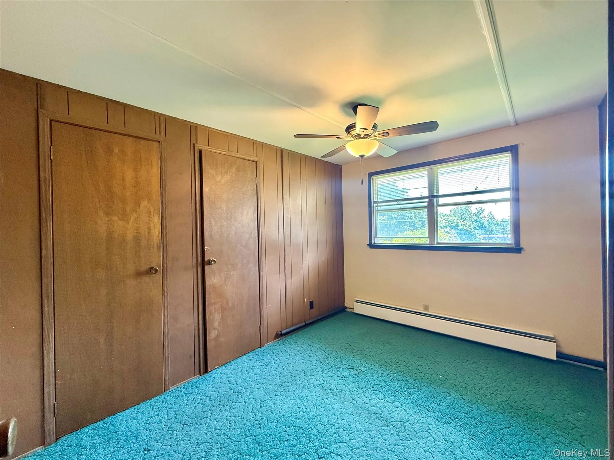Unfurnished bedroom with a baseboard radiator, carpet, wooden walls, and a ceiling fan Unfurnished bedroom with a baseboard radiator, carpet, wooden walls, and a ceiling fan