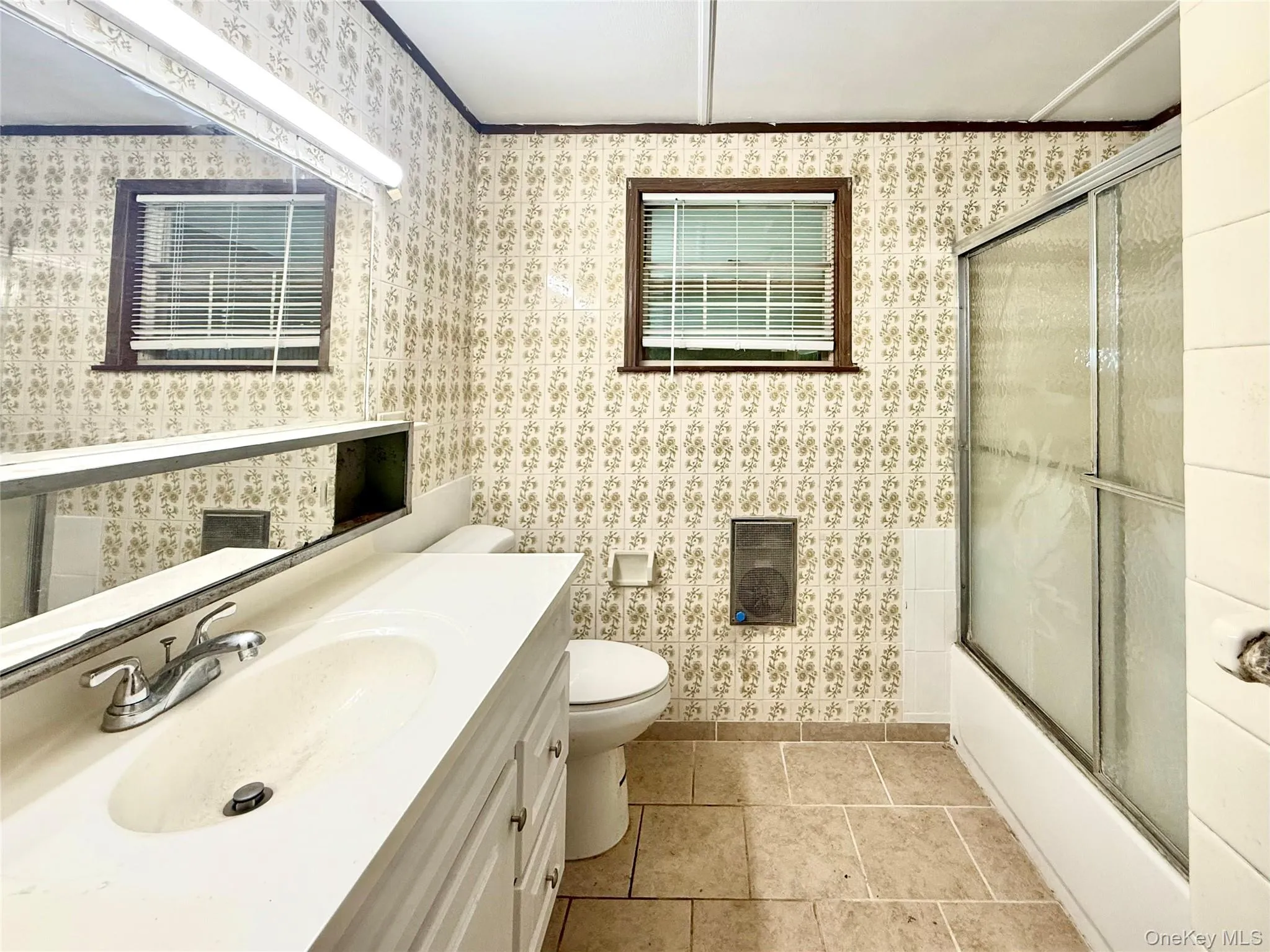 Bathroom with wallpapered walls, toilet, vanity, enclosed tub / shower combo, and baseboards Bathroom with wallpapered walls, toilet, vanity, enclosed tub / shower combo, and baseboards