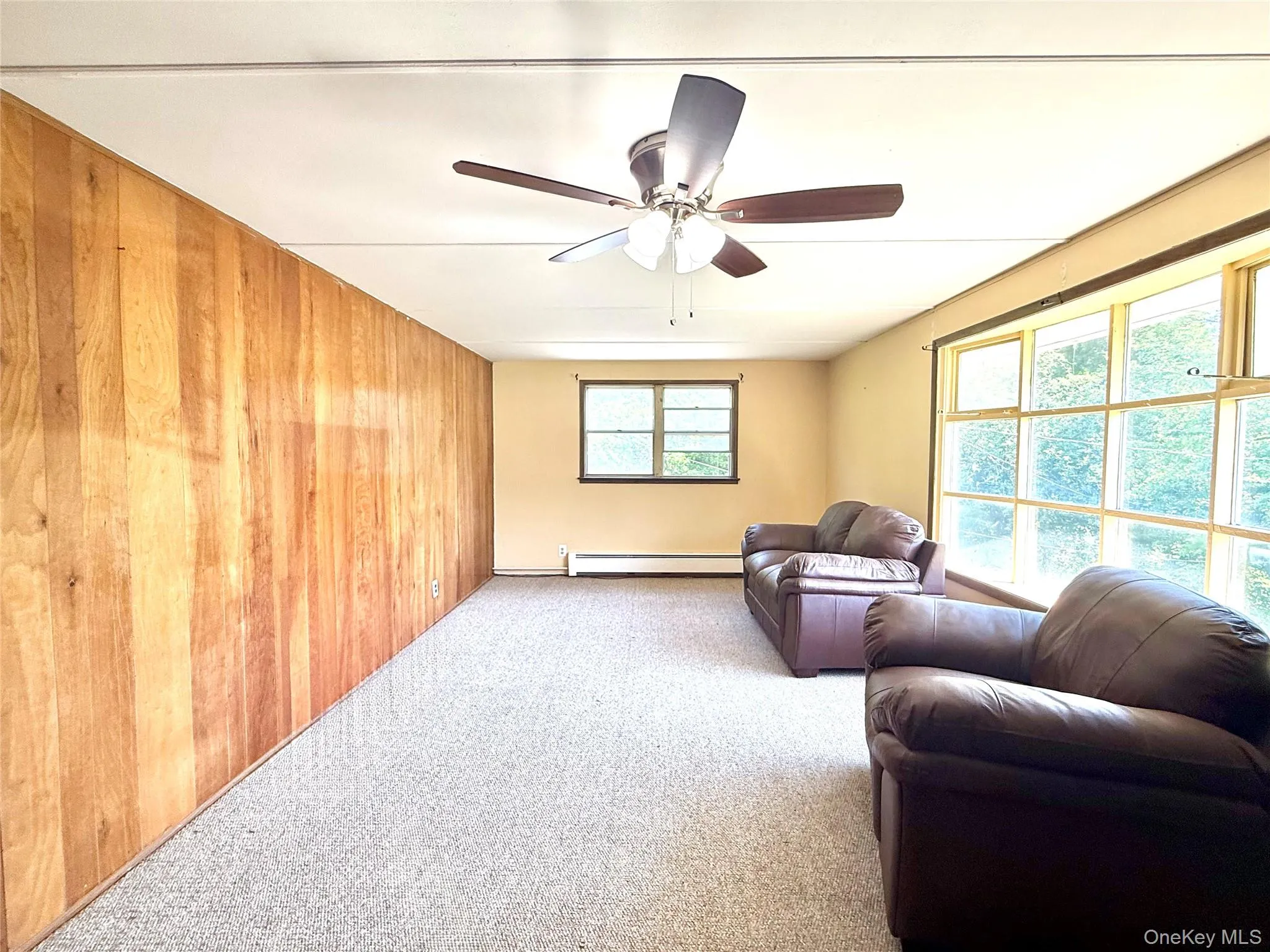Carpeted living room featuring baseboard heating, a ceiling fan, and wooden walls Carpeted living room featuring baseboard heating, a ceiling fan, and wooden walls