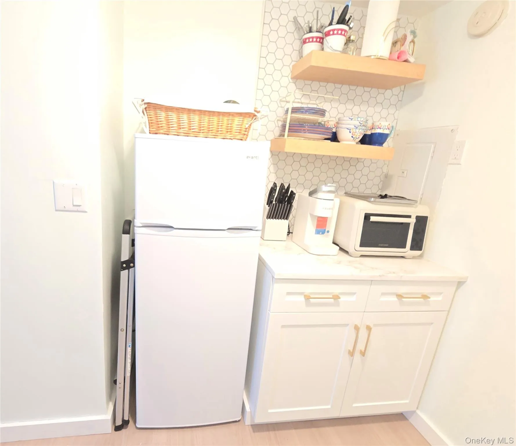 Compact kitchen area featuring a white refrigerator, white shaker-style cabinetry with gold-tone hardware, and a light-colored countertop Compact kitchen area featuring a white refrigerator, white shaker-style cabinetry with gold-tone hardware, and a light-colored countertop