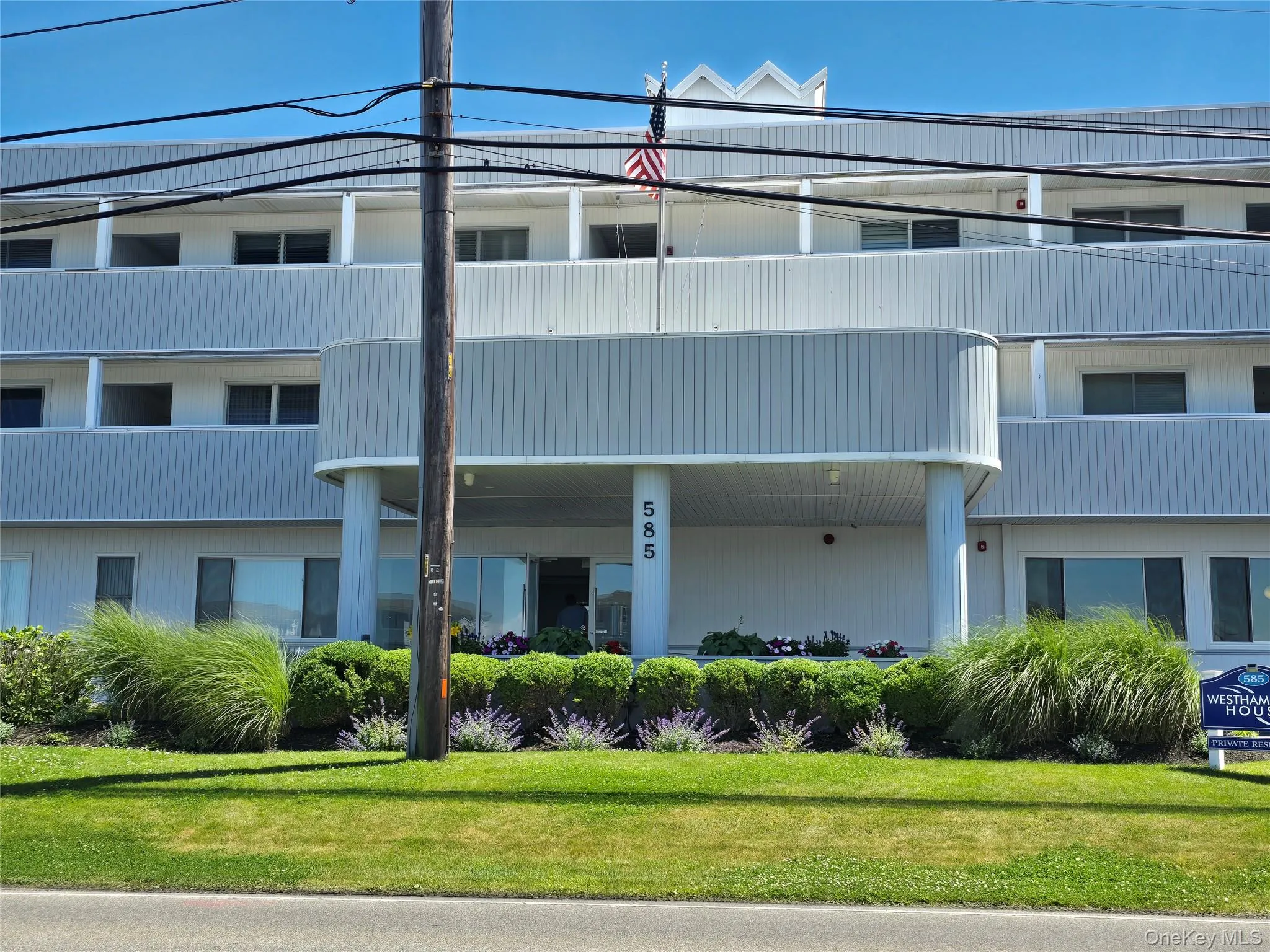 Multi-story building featuring a prominent covered entrance with columns, horizontal siding, multiple balconies, and a manicured landscape with green grass and various plantings Multi-story building featuring a prominent covered entrance with columns, horizontal siding, multiple balconies, and a manicured landscape with green grass and various plantings