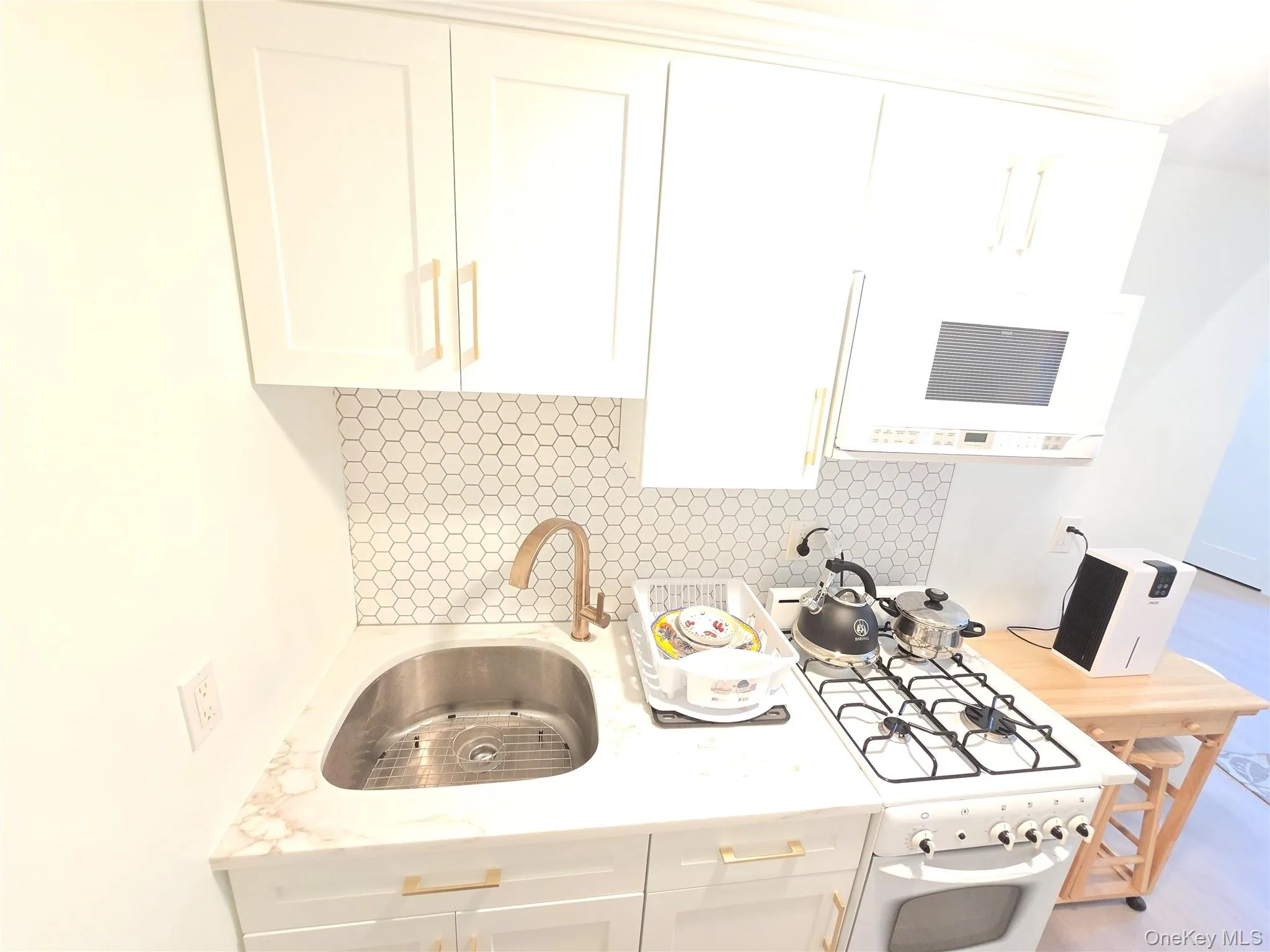 Compact kitchen featuring white shaker-style cabinetry with gold-tone hardware, a stainless steel undermount sink, a white gas range, and a white honeycomb tile backsplash Compact kitchen featuring white shaker-style cabinetry with gold-tone hardware, a stainless steel undermount sink, a white gas range, and a white honeycomb tile backsplash