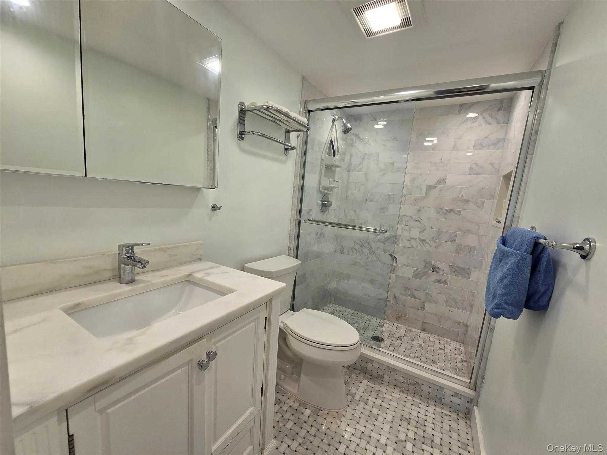 Bathroom featuring a single vanity with a white countertop and integrated sink, a mirrored medicine cabinet, a walk-in shower with a glass enclosure and marbled wall tiles, and mosaic tile flooring Bathroom featuring a single vanity with a white countertop and integrated sink, a mirrored medicine cabinet, a walk-in shower with a glass enclosure and marbled wall tiles, and mosaic tile flooring