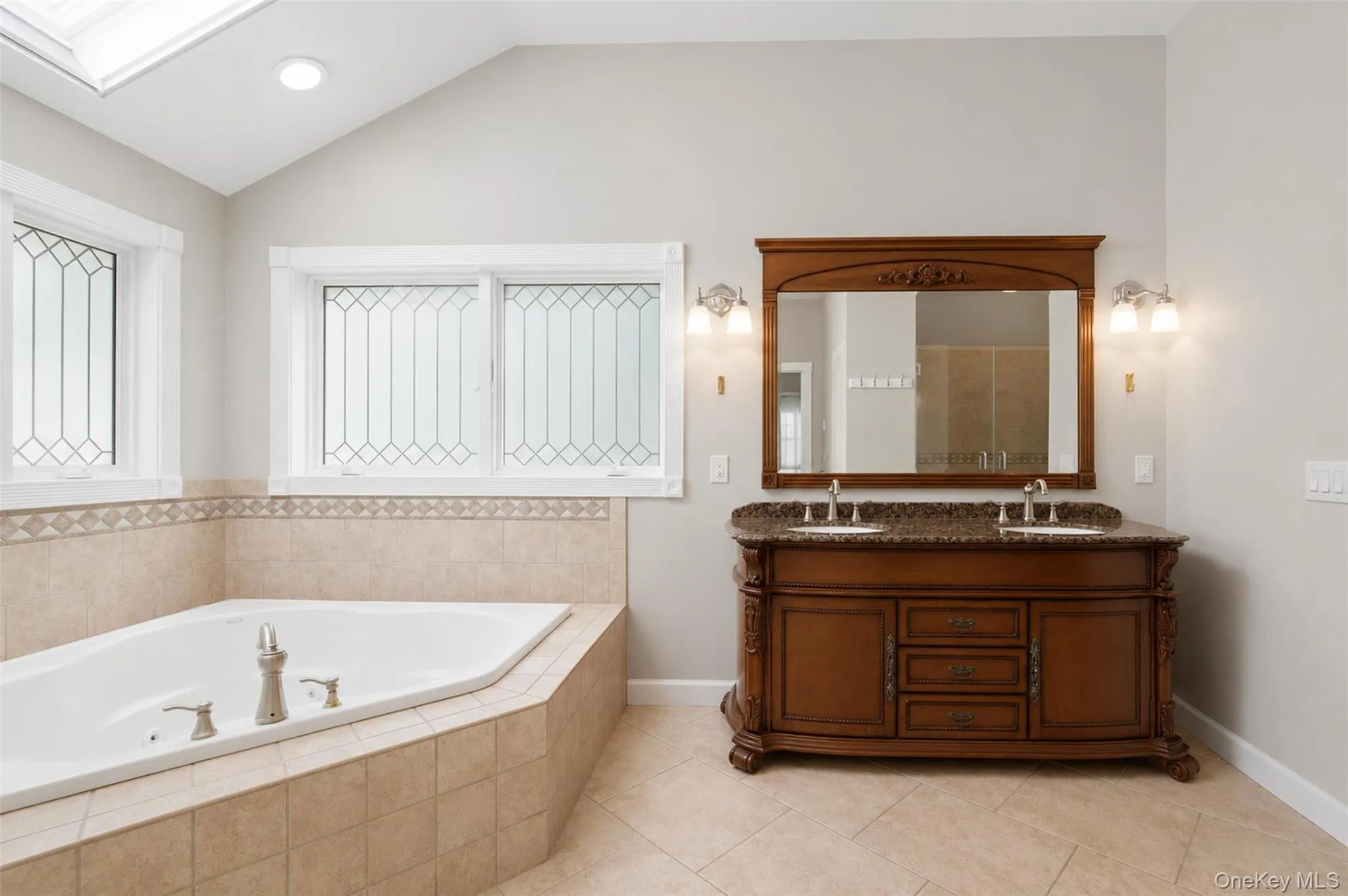 Soaking jet tub with tiled surround, dual-sink vanity with stone countertop, decorative windows, and vaulted ceiling with recessed lighting and skylight Soaking jet tub with tiled surround, dual-sink vanity with stone countertop, decorative windows, and vaulted ceiling with recessed lighting and skylight