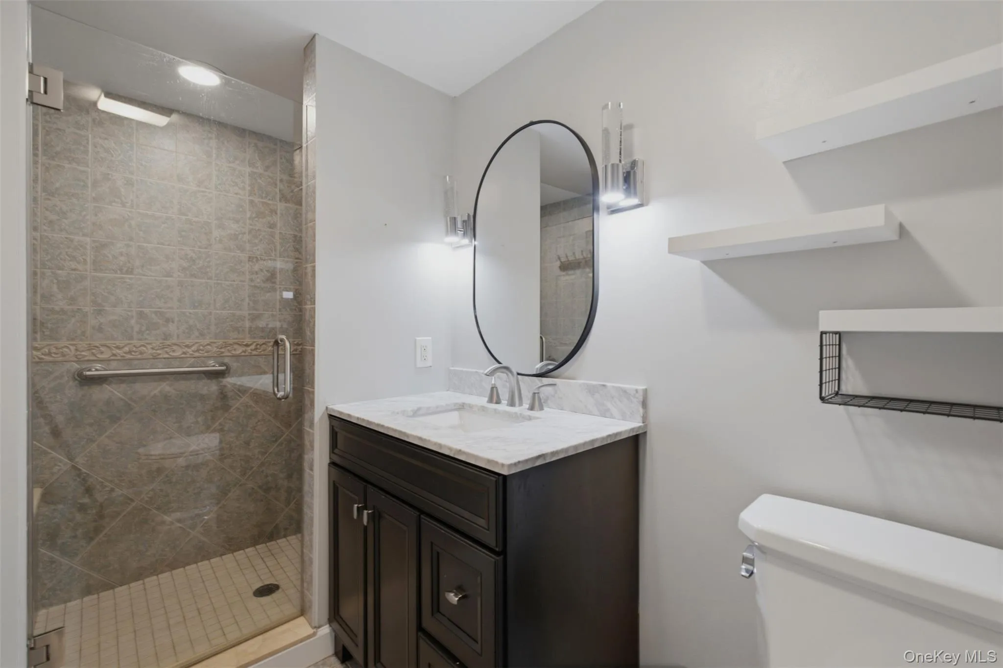 Bathroom featuring a glass-enclosed shower with tile surround, dark wood-finish vanity with a light-toned countertop, oval mirror, and wall-mounted shelving Bathroom featuring a glass-enclosed shower with tile surround, dark wood-finish vanity with a light-toned countertop, oval mirror, and wall-mounted shelving