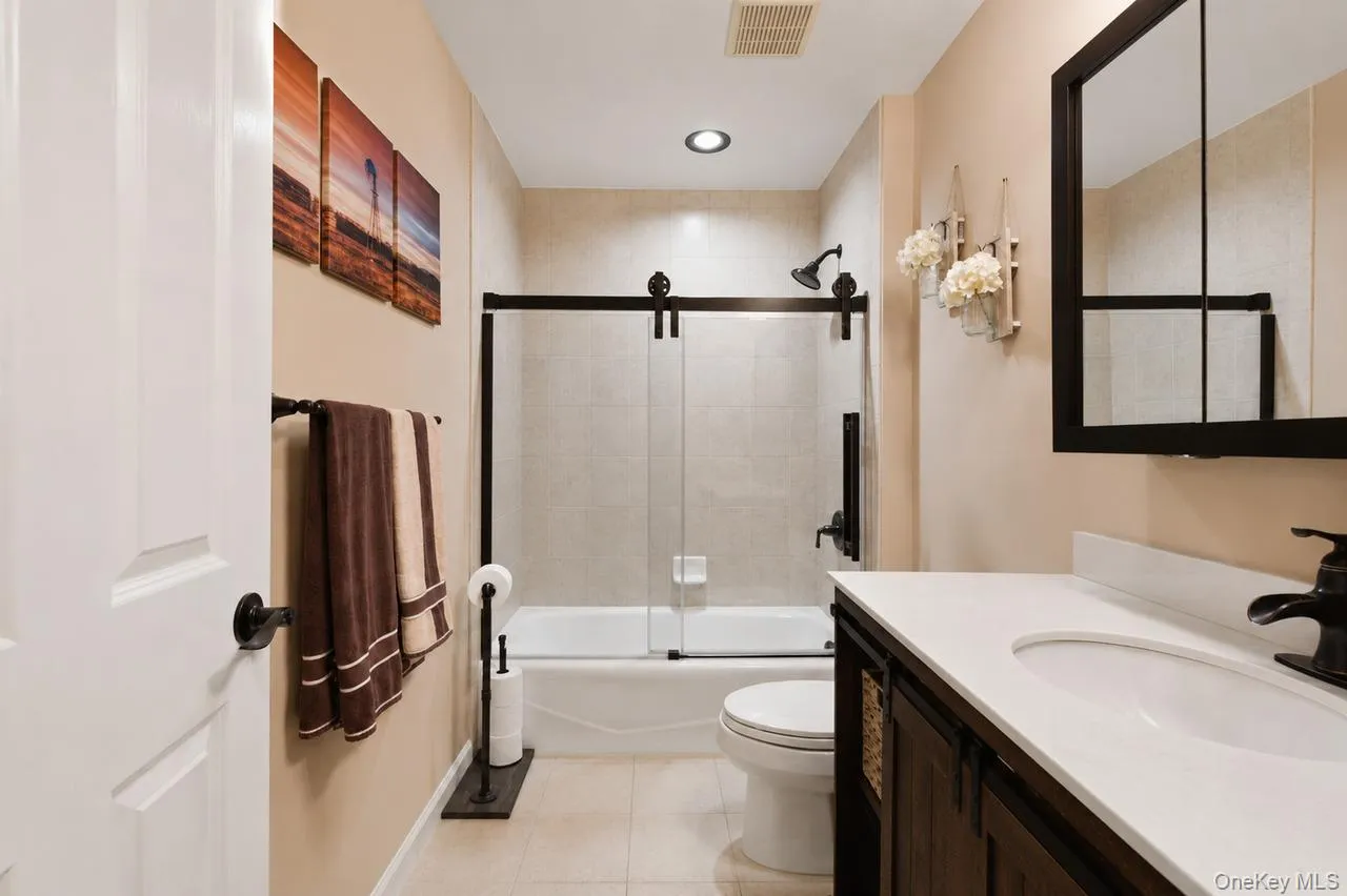Bathroom featuring a white bathtub with a sliding glass door and dark bronze hardware, white vanity with an integrated sink and dark bronze faucet, dark wood-finish vanity cabinetry, and recessed lighting Bathroom featuring a white bathtub with a sliding glass door and dark bronze hardware, white vanity with an integrated sink and dark bronze faucet, dark wood-finish vanity cabinetry, and recessed lighting