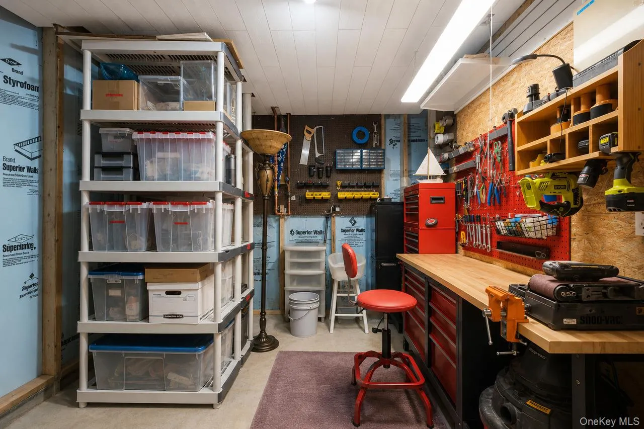 Utility room featuring extensive built-in shelving, a workbench with a light wood countertop, red storage drawers, a pegboard tool organization system, and overhead fluorescent lighting Utility room featuring extensive built-in shelving, a workbench with a light wood countertop, red storage drawers, a pegboard tool organization system, and overhead fluorescent lighting