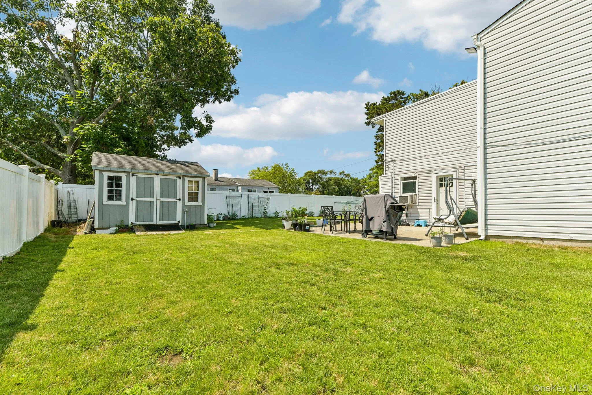 Expansive backyard featuring a manicured lawn, white vinyl privacy fencing, a gray storage shed with white trim, a concrete patio, and a light-colored siding exterior Expansive backyard featuring a manicured lawn, white vinyl privacy fencing, a gray storage shed with white trim, a concrete patio, and a light-colored siding exterior