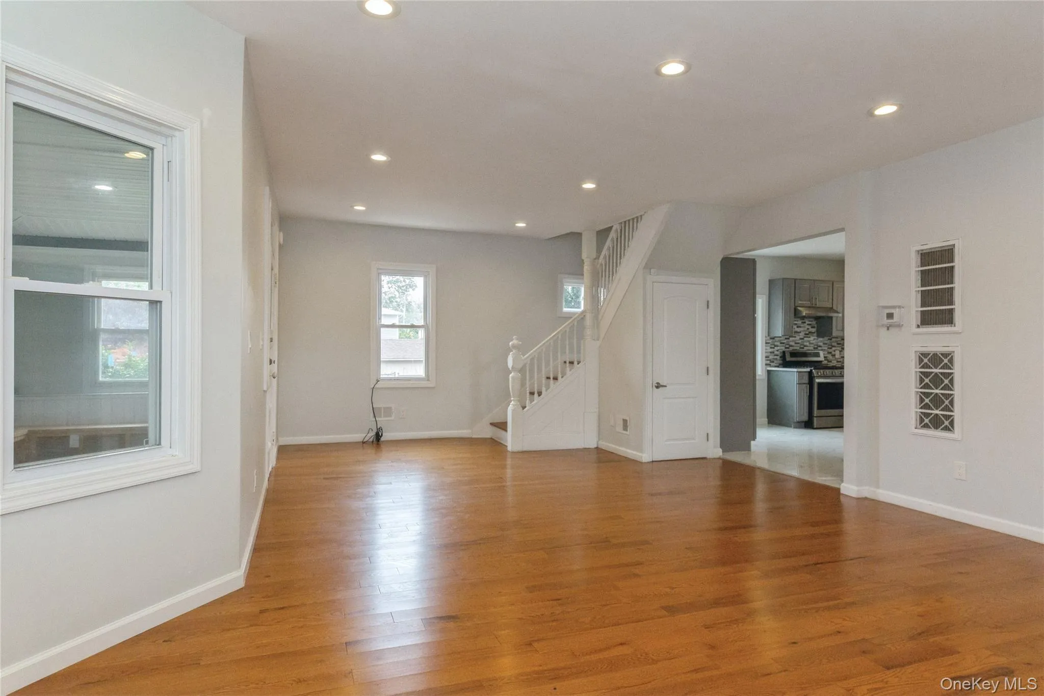 Expansive living area featuring warm wood-finish flooring, recessed lighting, white trim, and a staircase with white balusters Expansive living area featuring warm wood-finish flooring, recessed lighting, white trim, and a staircase with white balusters