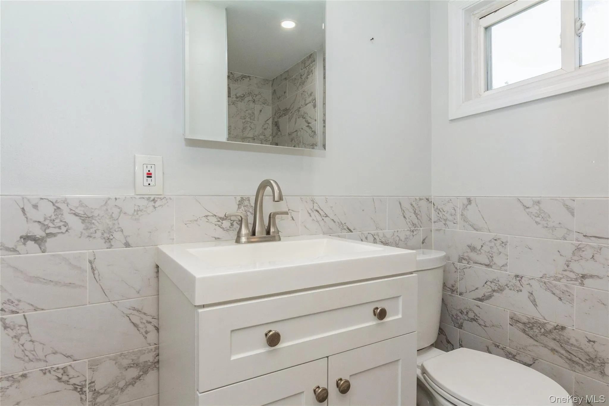 White vanity with integrated sink, brushed nickel faucet, wall-mounted mirror, and white tiled half-wall with gray veining White vanity with integrated sink, brushed nickel faucet, wall-mounted mirror, and white tiled half-wall with gray veining