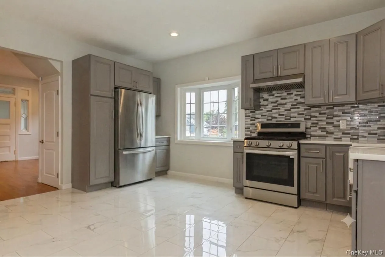 Kitchen featuring white marble-finish tile flooring, grey cabinetry, stainless steel appliances, a multi-tone tile backsplash, and a bay window Kitchen featuring white marble-finish tile flooring, grey cabinetry, stainless steel appliances, a multi-tone tile backsplash, and a bay window