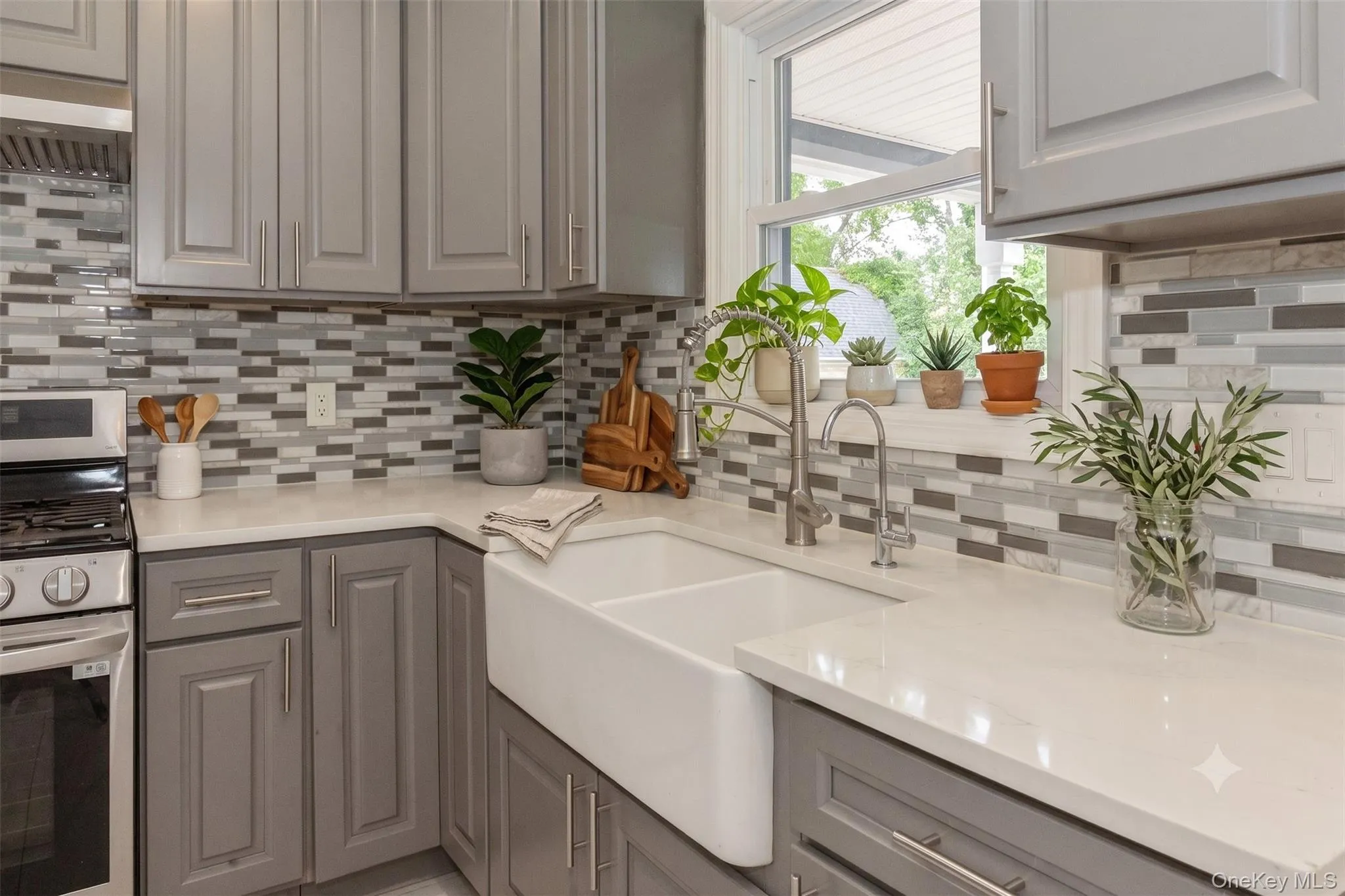 Modern kitchen featuring a white farmhouse-style sink, light gray cabinetry, stainless steel appliances, a mosaic tile backsplash, and light-colored countertops Modern kitchen featuring a white farmhouse-style sink, light gray cabinetry, stainless steel appliances, a mosaic tile backsplash, and light-colored countertops