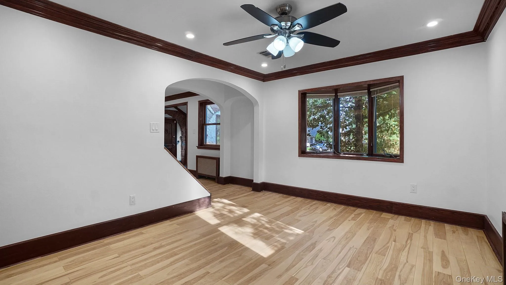 Expansive room featuring wood-finish flooring, a bay window with dark trim, and a ceiling fan with integrated lighting Expansive room featuring wood-finish flooring, a bay window with dark trim, and a ceiling fan with integrated lighting