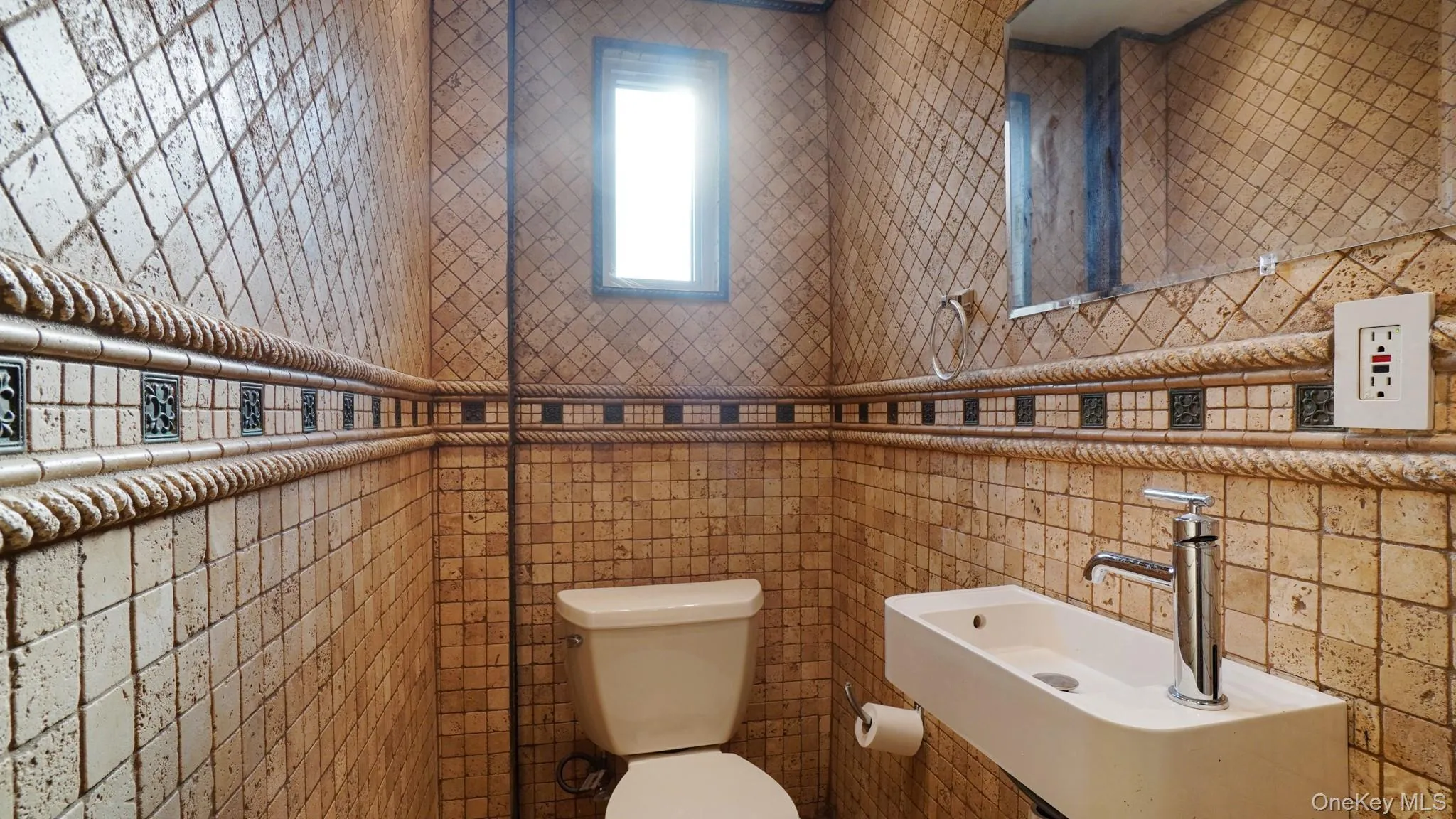 Powder room featuring full-height mosaic-style tiling with decorative trim, a modern wall-mounted sink with a chrome faucet, a rectangular mirror, and a small window Powder room featuring full-height mosaic-style tiling with decorative trim, a modern wall-mounted sink with a chrome faucet, a rectangular mirror, and a small window