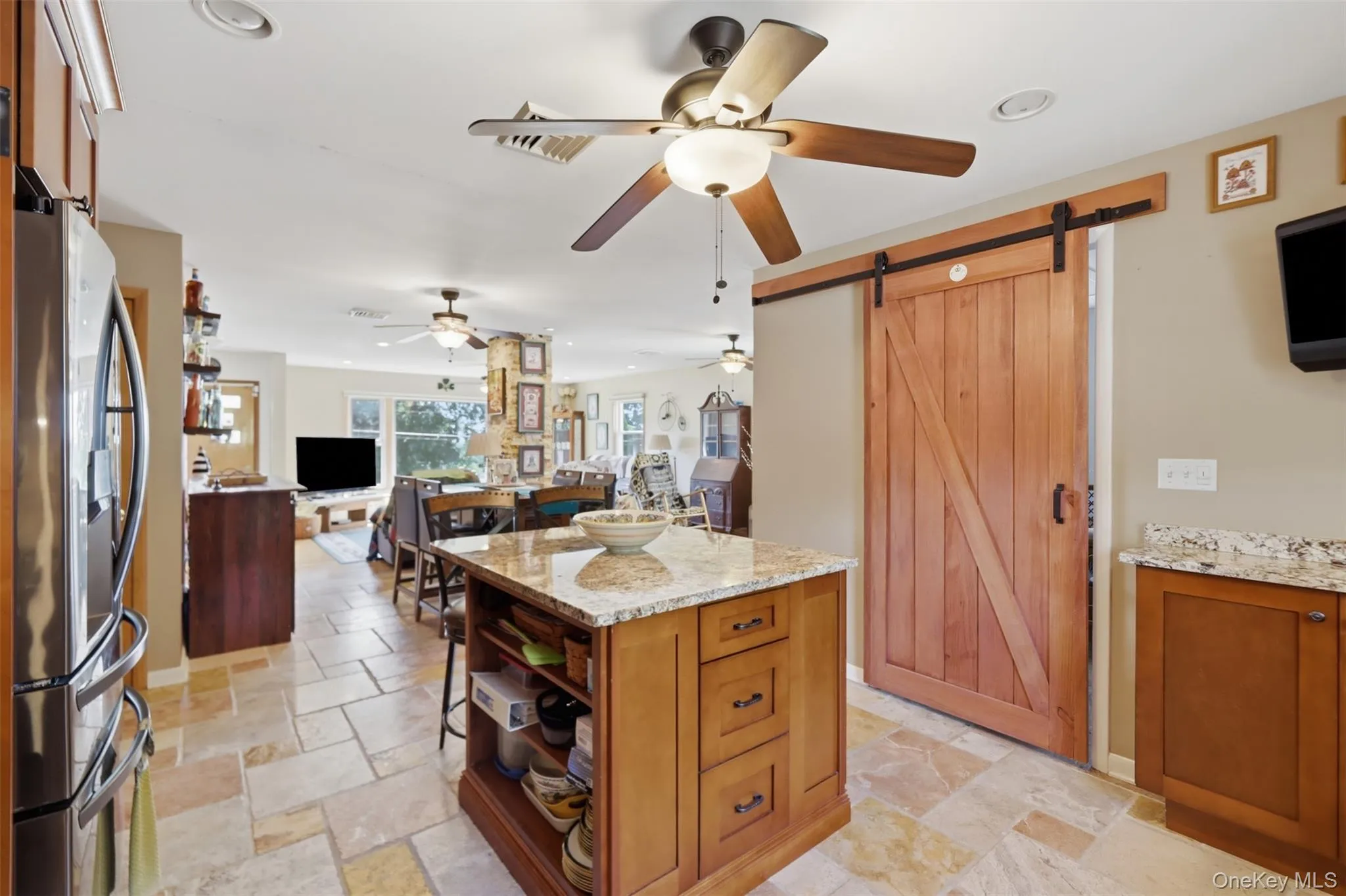 Kitchen featuring a central island with a granite countertop and storage, stainless steel refrigerator, light-toned stone tile flooring, wood-paneled barn door, and recessed lighting Kitchen featuring a central island with a granite countertop and storage, stainless steel refrigerator, light-toned stone tile flooring, wood-paneled barn door, and recessed lighting