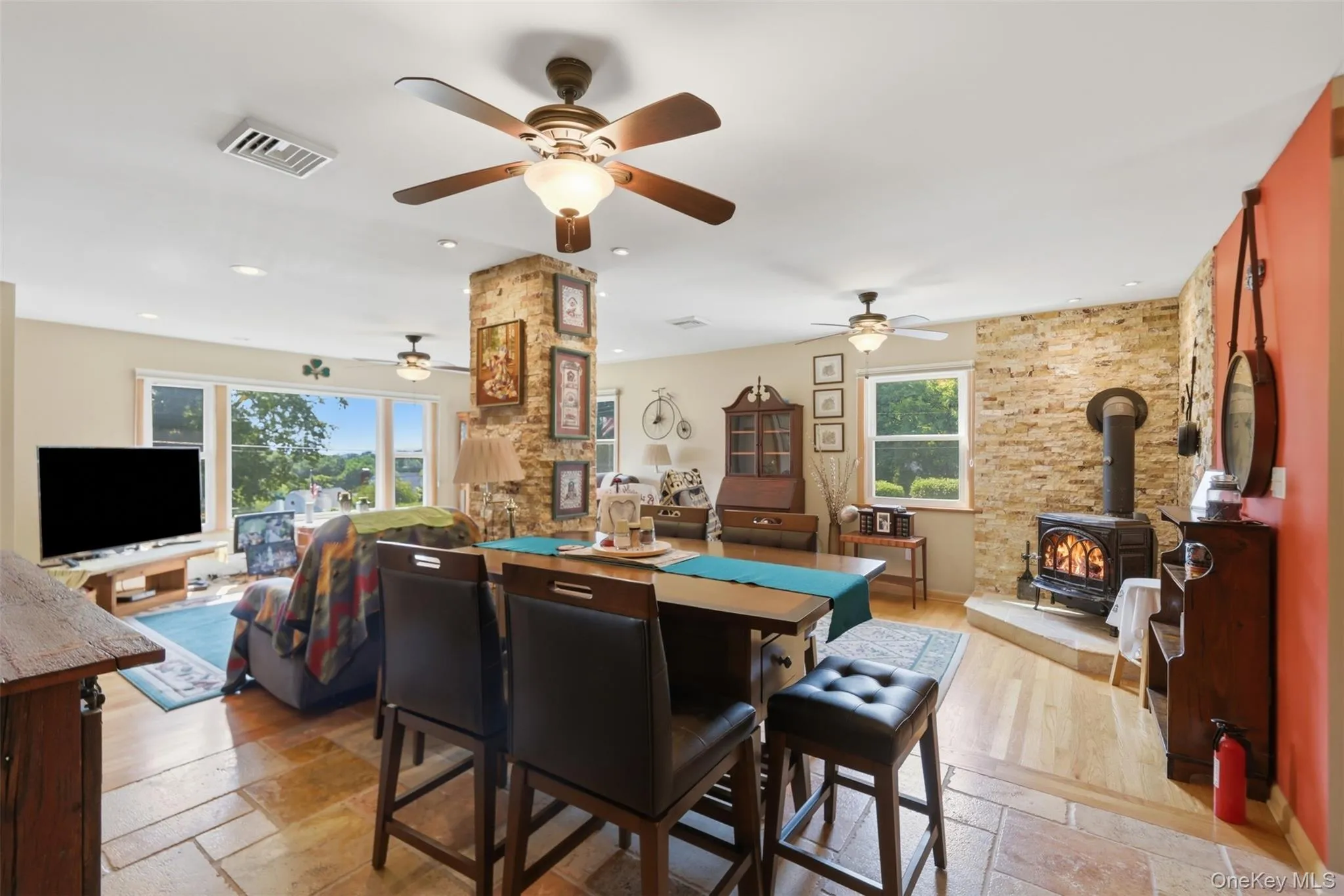 Open concept living space featuring stone-finish flooring, a stone-clad structural column, and a prominent wood-burning stove with a stone hearth Open concept living space featuring stone-finish flooring, a stone-clad structural column, and a prominent wood-burning stove with a stone hearth