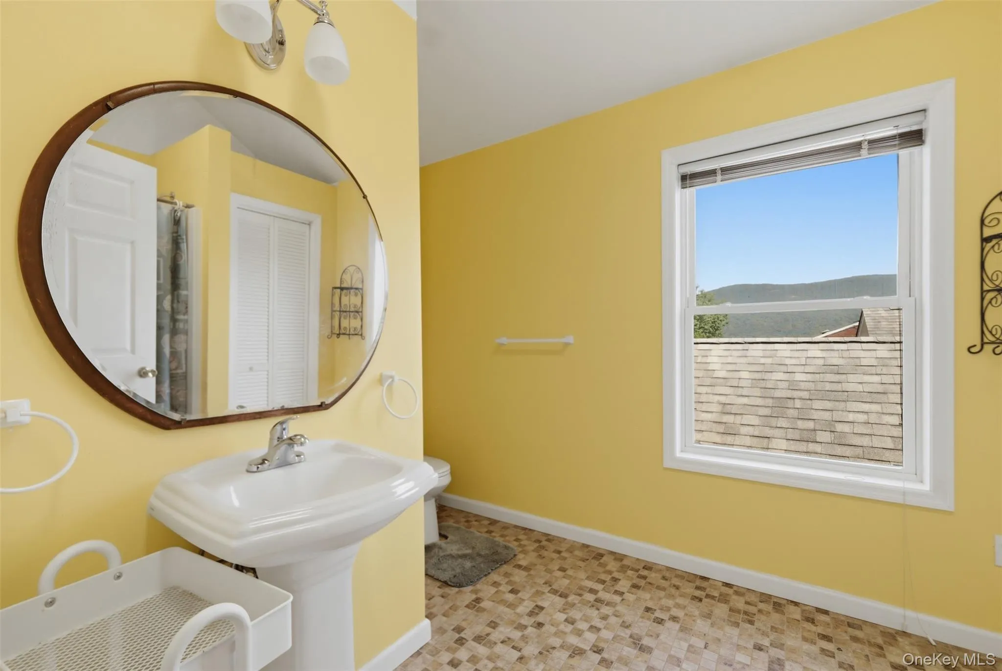 Bathroom featuring a pedestal sink, oval mirror with wood-finish frame, and window with a mountain backdrop Bathroom featuring a pedestal sink, oval mirror with wood-finish frame, and window with a mountain backdrop