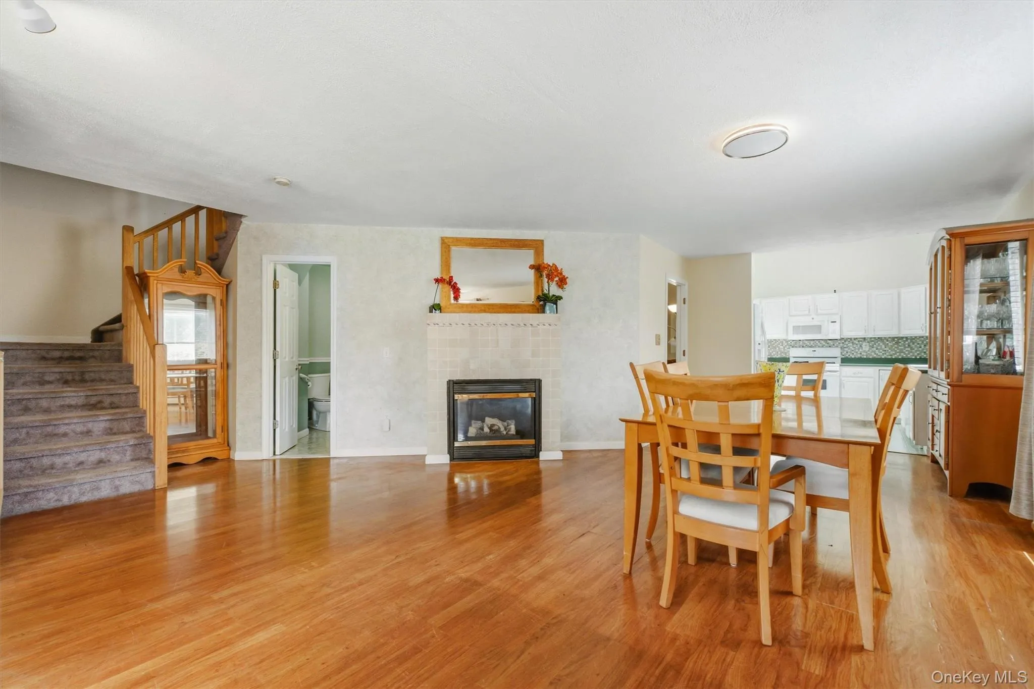 Open-concept living space with wood-finish flooring, a central fireplace featuring a tiled surround and wood mantel, and a visible staircase with carpeted treads and wood railings Open-concept living space with wood-finish flooring, a central fireplace featuring a tiled surround and wood mantel, and a visible staircase with carpeted treads and wood railings
