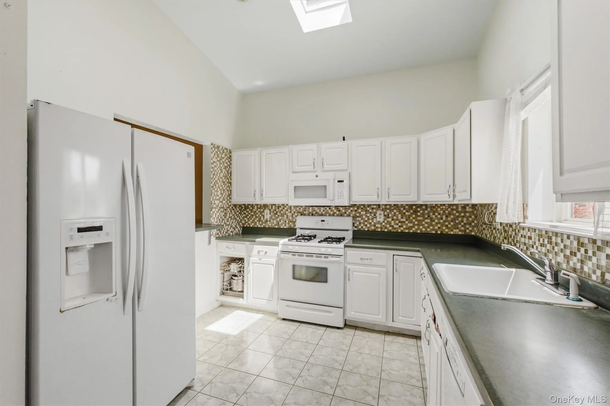 Kitchen featuring white cabinetry, white appliances, a mosaic tile backsplash, dark countertops, and tile flooring Kitchen featuring white cabinetry, white appliances, a mosaic tile backsplash, dark countertops, and tile flooring