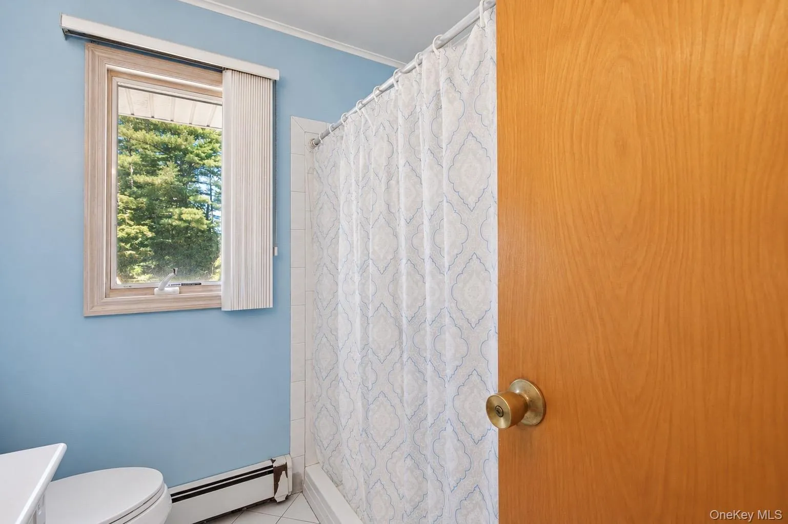 Bathroom featuring light blue wall paint, a window with natural wood trim, white tile flooring, and a shower with white subway tile surround Bathroom featuring light blue wall paint, a window with natural wood trim, white tile flooring, and a shower with white subway tile surround