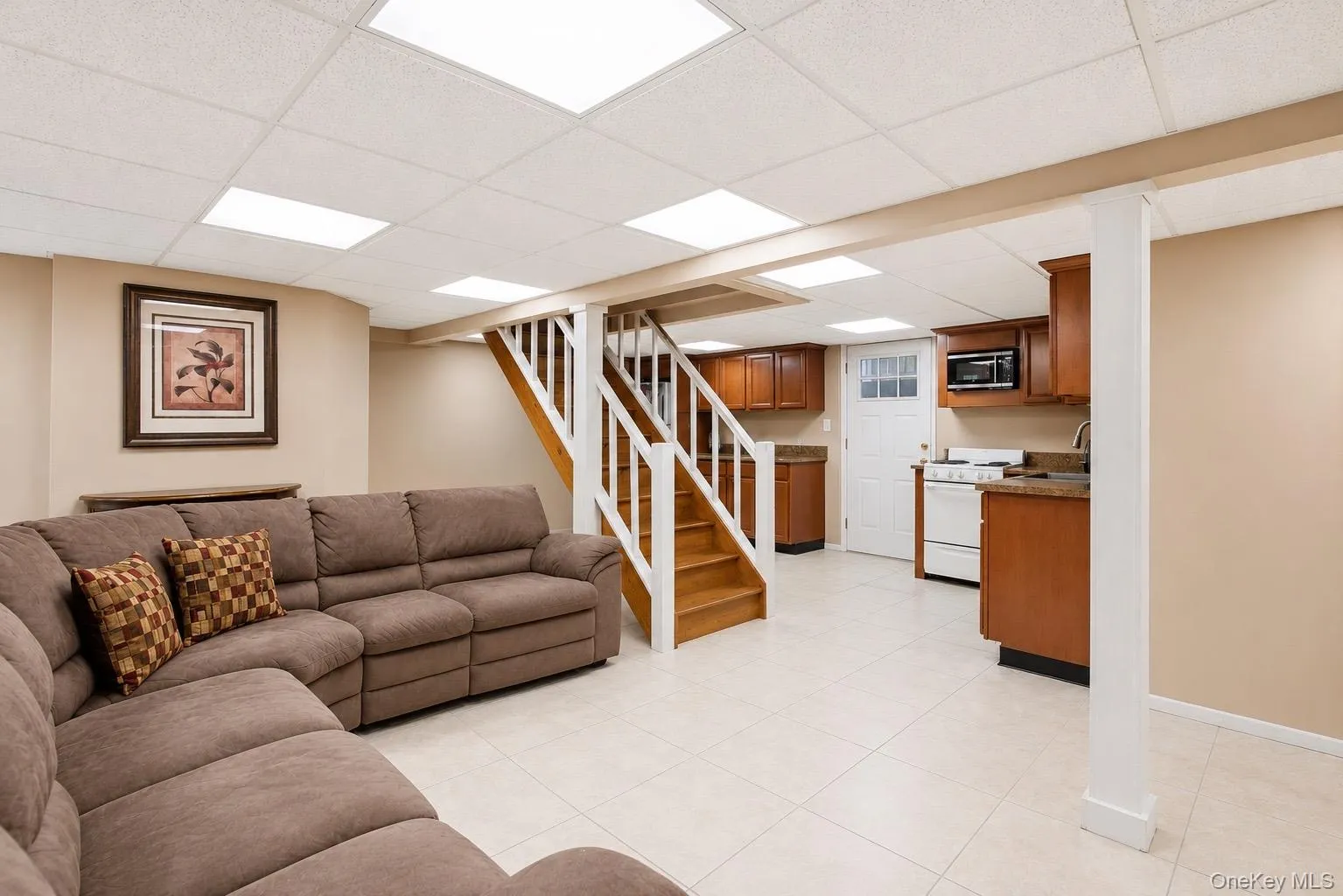 Finished basement featuring a full kitchen, wood-finish staircase with white risers, tiled flooring, drop ceiling with recessed lighting, and a neutral color palette open floor plan OSE Finished basement featuring a full kitchen, wood-finish staircase with white risers, tiled flooring, drop ceiling with recessed lighting, and a neutral color palette open floor plan OSE