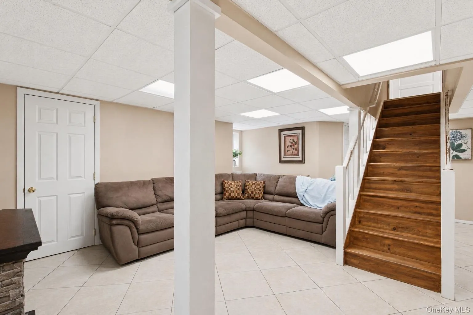 Spacious room featuring light-toned tile flooring, a drop ceiling with recessed lighting, and a prominent wood staircase with white risers and railings Spacious room featuring light-toned tile flooring, a drop ceiling with recessed lighting, and a prominent wood staircase with white risers and railings