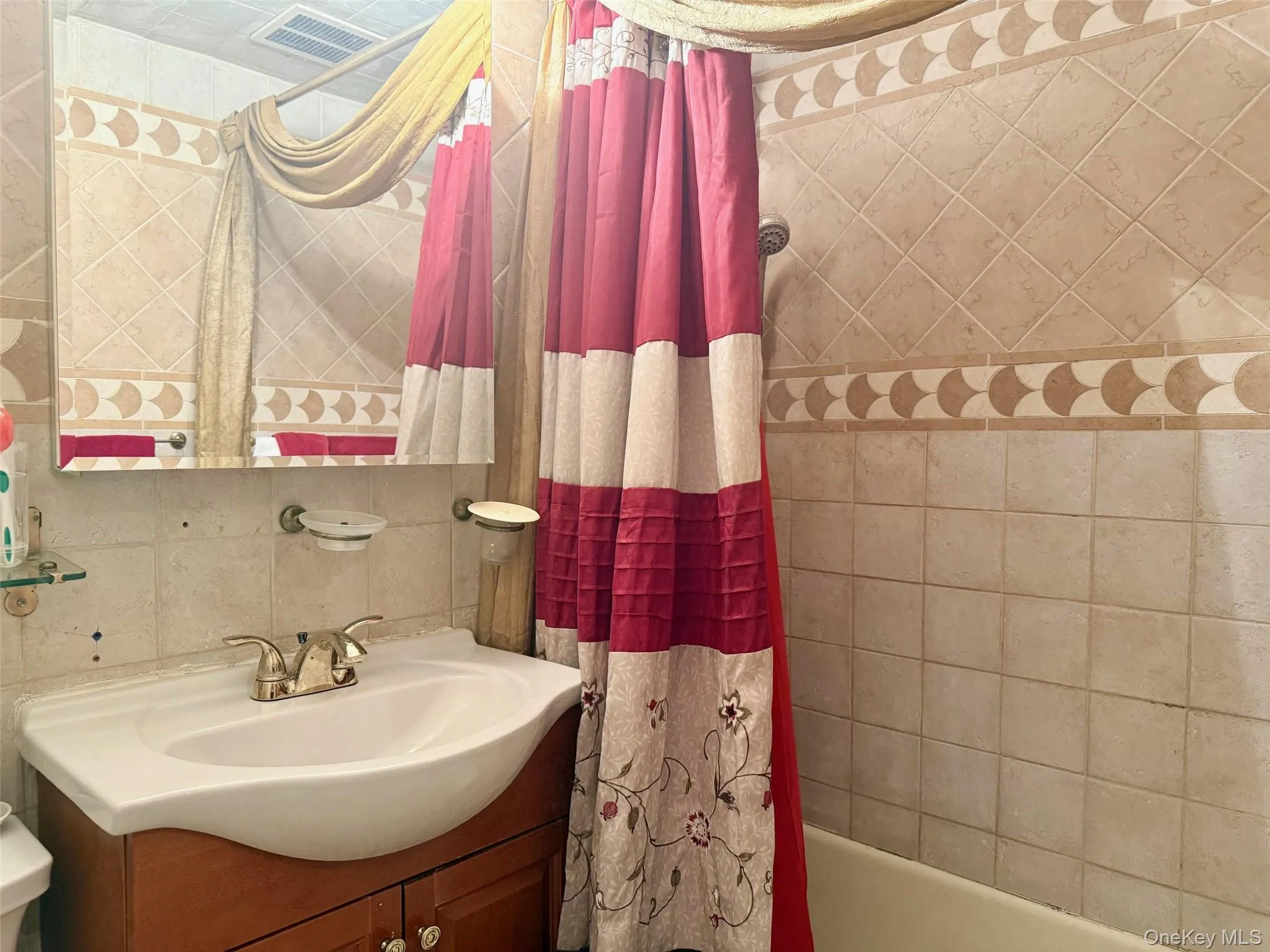 Bathroom featuring a tiled shower surround with decorative accent banding, a wood-finish vanity with an integrated sink, a wall-mounted mirror, and a shower/tub combination Bathroom featuring a tiled shower surround with decorative accent banding, a wood-finish vanity with an integrated sink, a wall-mounted mirror, and a shower/tub combination