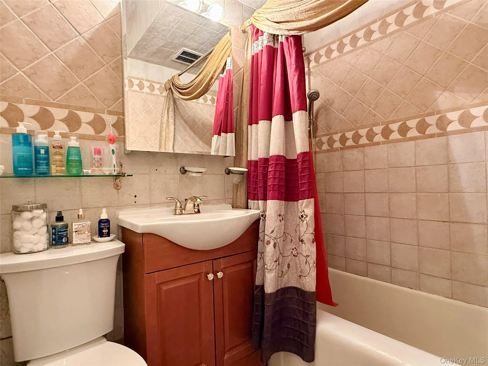 Bathroom featuring a tiled shower surround with decorative accent banding, a wood-finish vanity with an integrated sink, a wall-mounted mirror, and a shower/tub combination Bathroom featuring a tiled shower surround with decorative accent banding, a wood-finish vanity with an integrated sink, a wall-mounted mirror, and a shower/tub combination