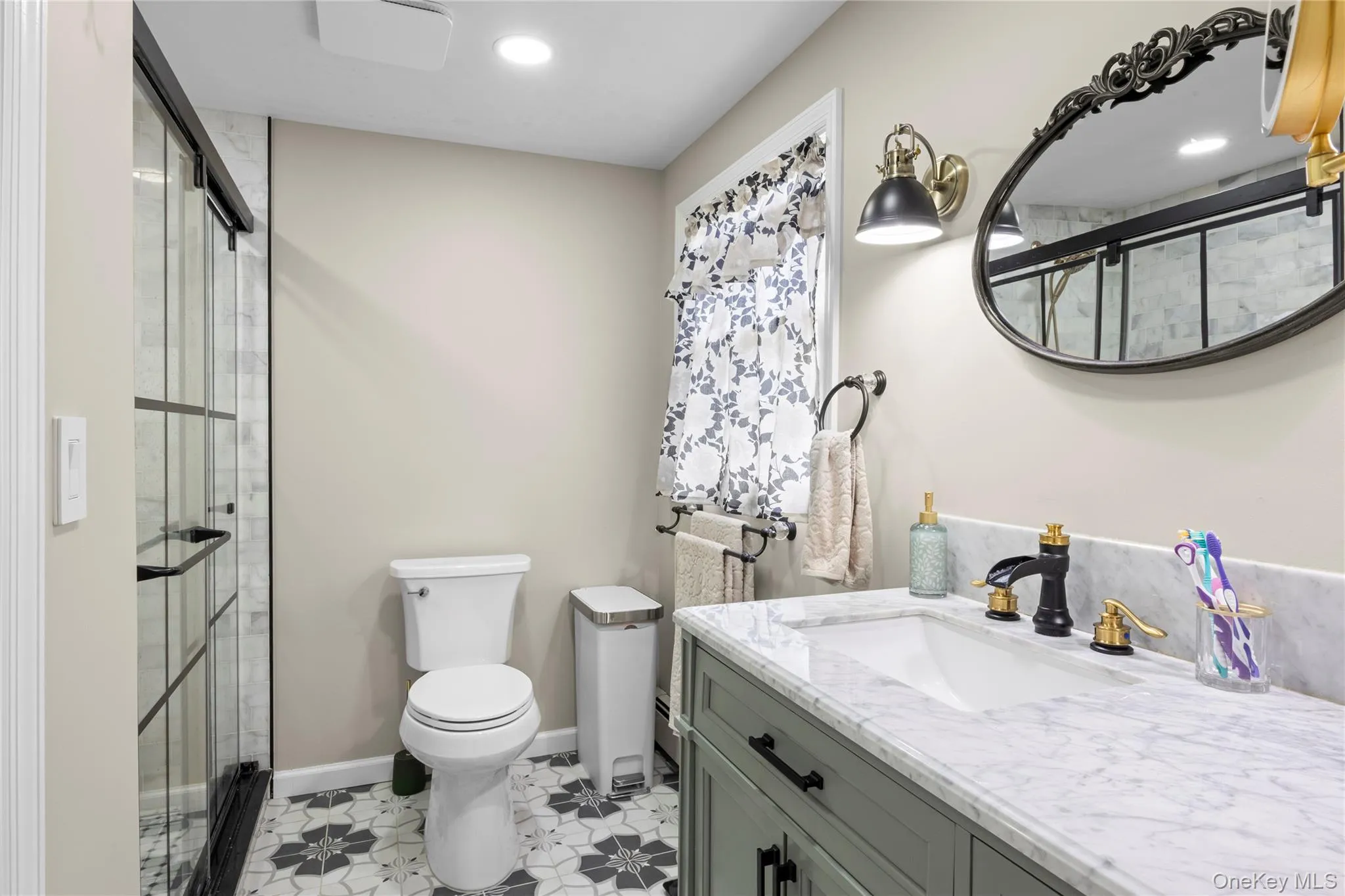Bathroom featuring a walk-in shower with black-framed glass enclosure, patterned floor tiling, single vanity with a marble-finish countertop, and decorative mirror Bathroom featuring a walk-in shower with black-framed glass enclosure, patterned floor tiling, single vanity with a marble-finish countertop, and decorative mirror