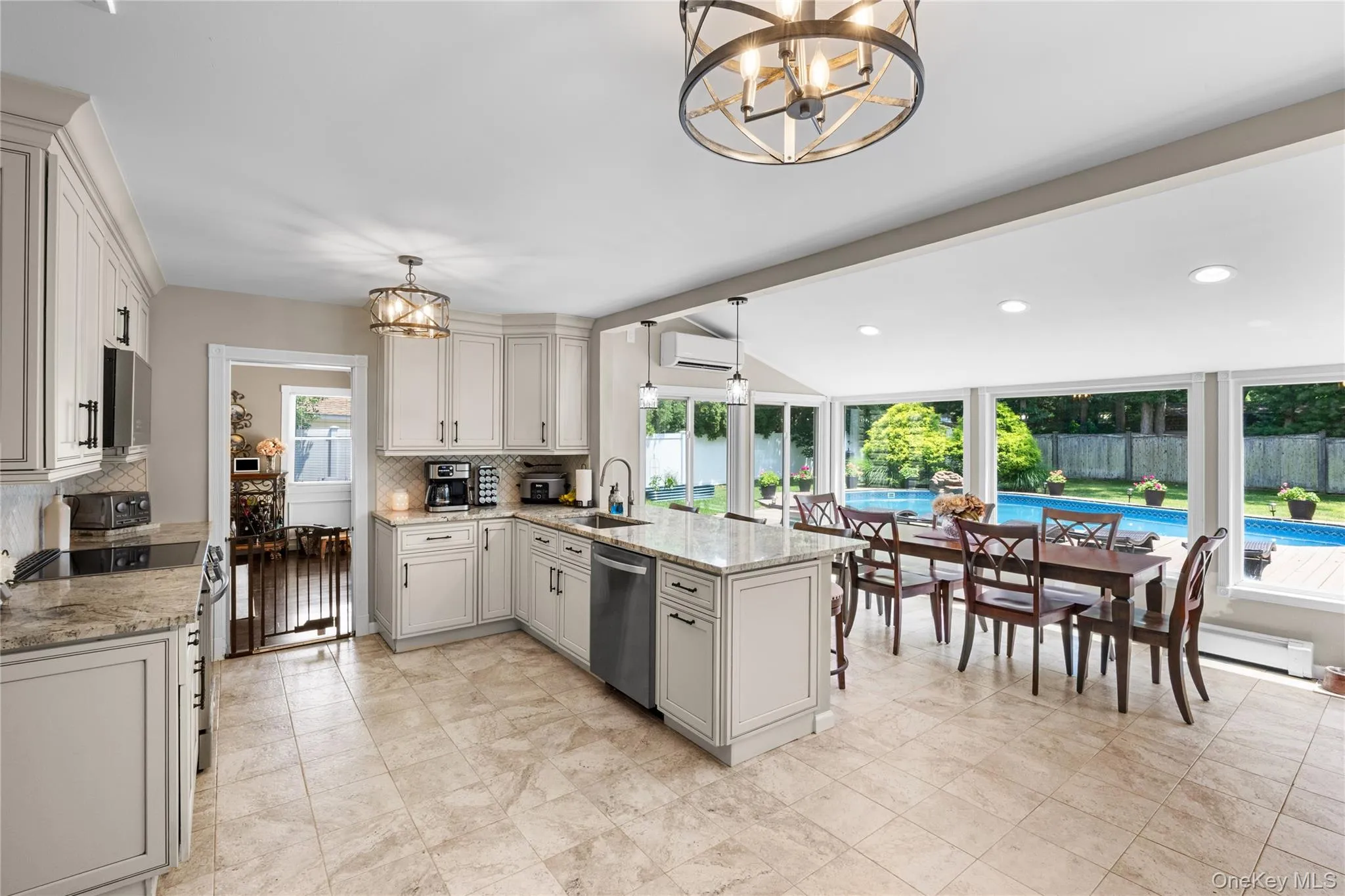 Kitchen and dining area featuring light cabinetry with dark hardware, granite countertops, a central island with a sink, tiled flooring, and a dining space with expansive windows overlooking a swimming pool Kitchen and dining area featuring light cabinetry with dark hardware, granite countertops, a central island with a sink, tiled flooring, and a dining space with expansive windows overlooking a swimming pool