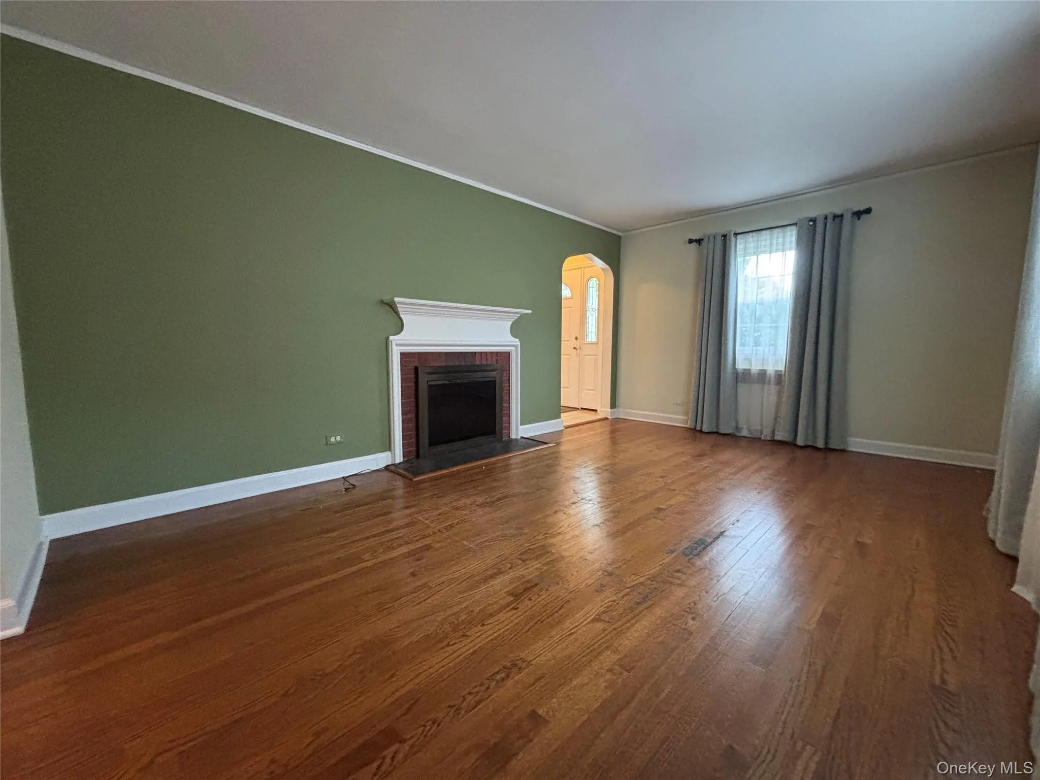 Spacious living area featuring wood-finish flooring, a fireplace with a white mantel and dark brick surround, and an arched entry Spacious living area featuring wood-finish flooring, a fireplace with a white mantel and dark brick surround, and an arched entry