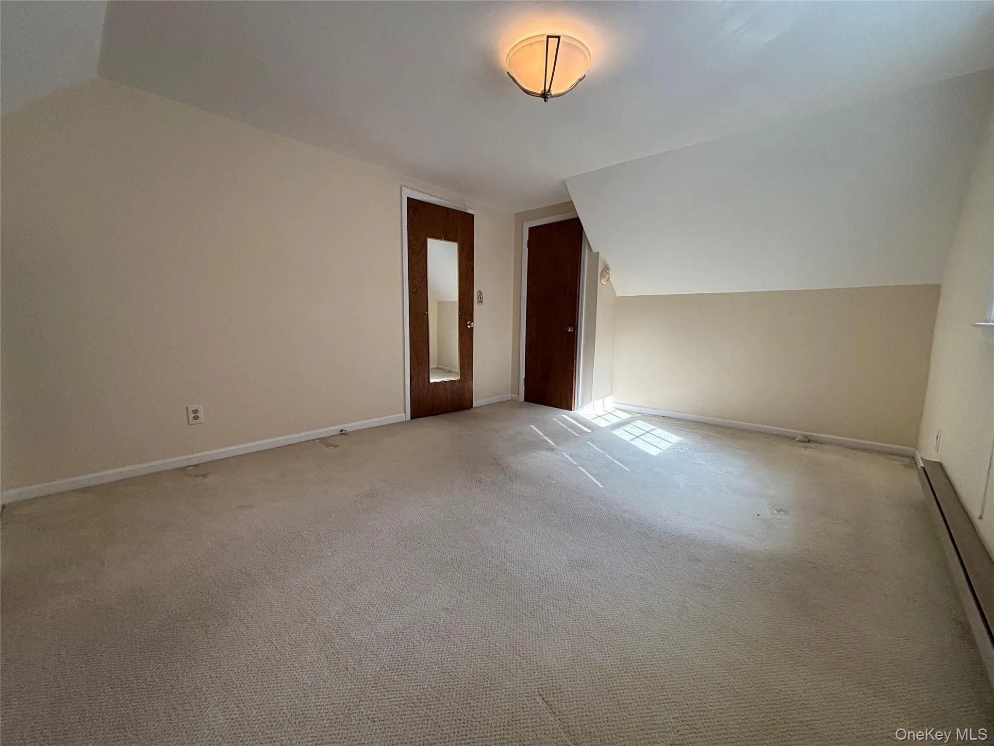 Carpeted room featuring a ceiling-mounted light fixture, two wood-finish doors, and an angled ceiling line Carpeted room featuring a ceiling-mounted light fixture, two wood-finish doors, and an angled ceiling line