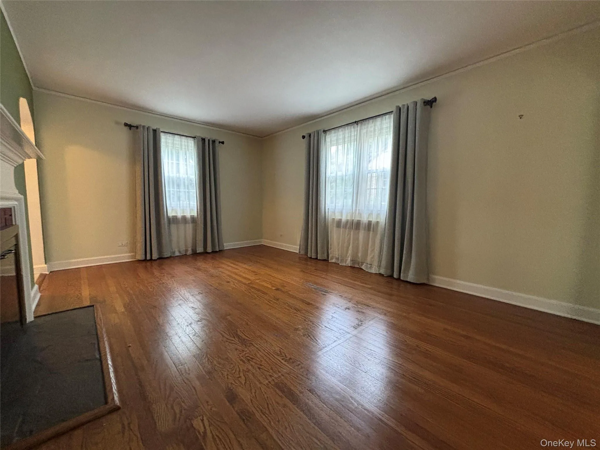 Spacious room featuring rich wood-finish flooring, two large windows, baseboard molding, crown molding, and a fireplace with a white mantel and dark hearth Spacious room featuring rich wood-finish flooring, two large windows, baseboard molding, crown molding, and a fireplace with a white mantel and dark hearth