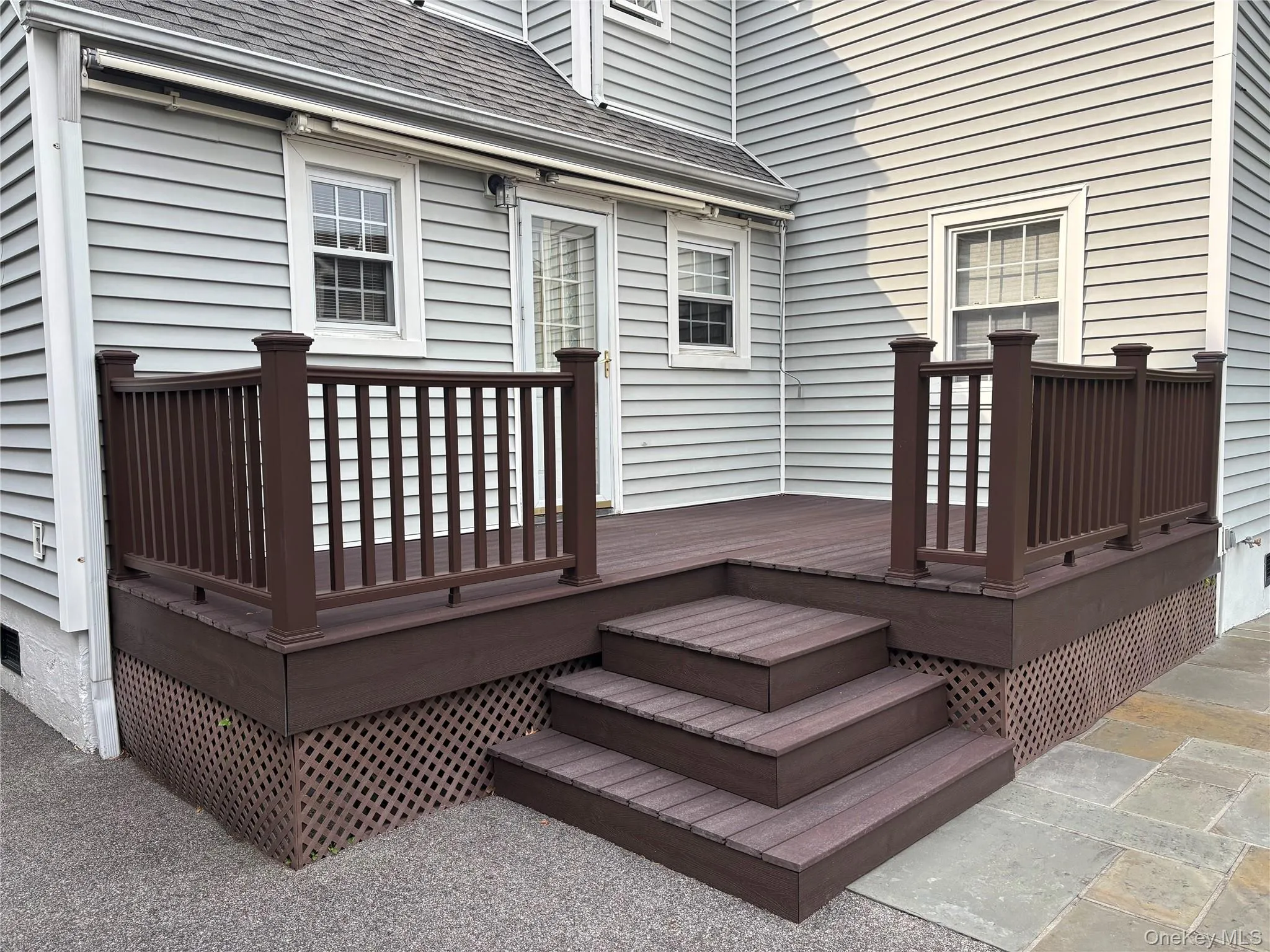 Multi-level composite deck featuring dark brown railing, tiered steps, lattice skirting, and stone patio surround Multi-level composite deck featuring dark brown railing, tiered steps, lattice skirting, and stone patio surround