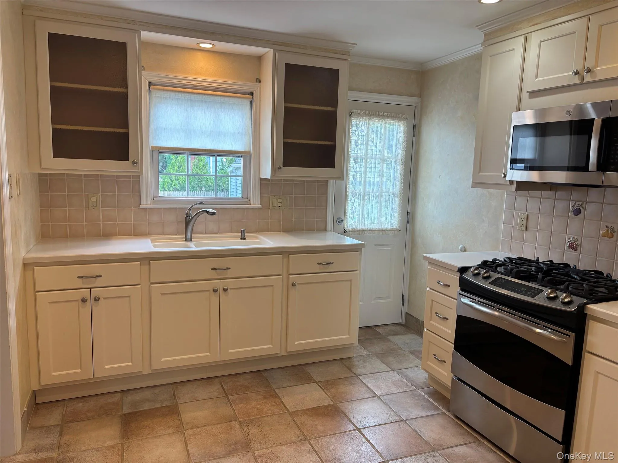 Kitchen featuring cream-colored cabinetry, a stainless steel microwave, a stainless steel range, light-toned countertops, and square floor tiles Kitchen featuring cream-colored cabinetry, a stainless steel microwave, a stainless steel range, light-toned countertops, and square floor tiles