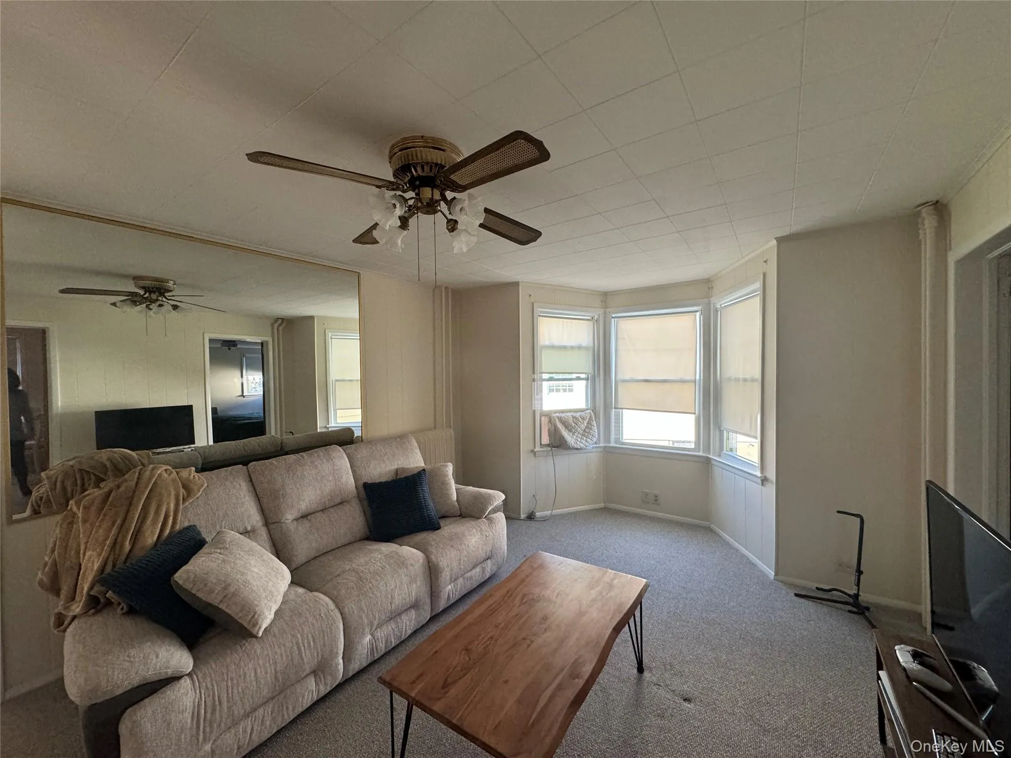 Living area featuring a bay window with multiple panes, textured carpeting, a ceiling fan with integrated lighting, and a mirrored wall accent Living area featuring a bay window with multiple panes, textured carpeting, a ceiling fan with integrated lighting, and a mirrored wall accent