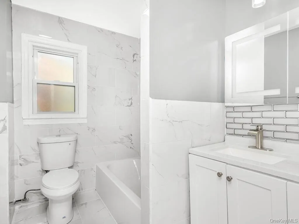 Bathroom featuring white tile flooring, white subway tile accents, a vanity with a white countertop and basin sink, a wall-mounted mirror, and a white toilet Bathroom featuring white tile flooring, white subway tile accents, a vanity with a white countertop and basin sink, a wall-mounted mirror, and a white toilet