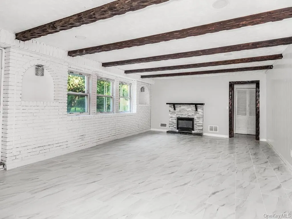 Spacious room featuring white painted brick walls with arched recesses, exposed wood ceiling beams, three windows with exterior views, light-toned tile flooring, and a stone-clad fireplace with a dark mantel Spacious room featuring white painted brick walls with arched recesses, exposed wood ceiling beams, three windows with exterior views, light-toned tile flooring, and a stone-clad fireplace with a dark mantel