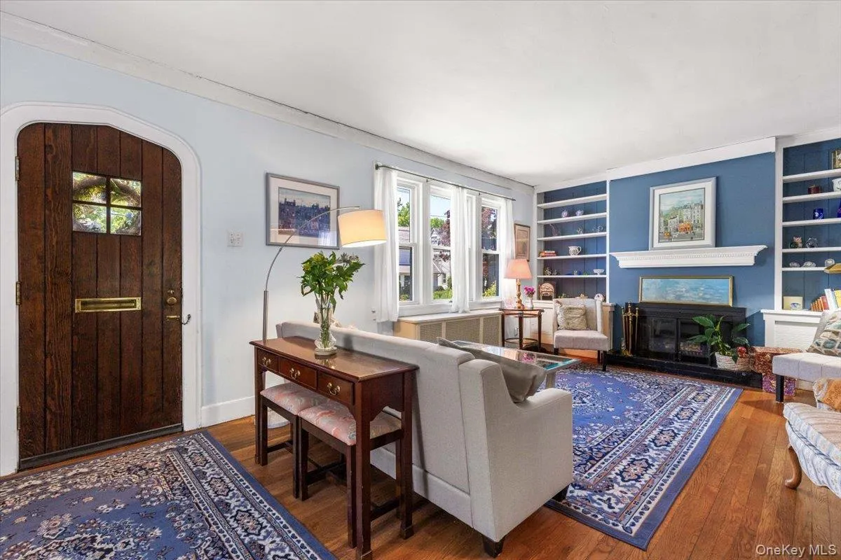 Hardwood flooring throughout this living space featuring a dark wood arched entry door with glass pane, a fireplace with a white mantel, and built-in shelving on a blue accent wall Hardwood flooring throughout this living space featuring a dark wood arched entry door with glass pane, a fireplace with a white mantel, and built-in shelving on a blue accent wall