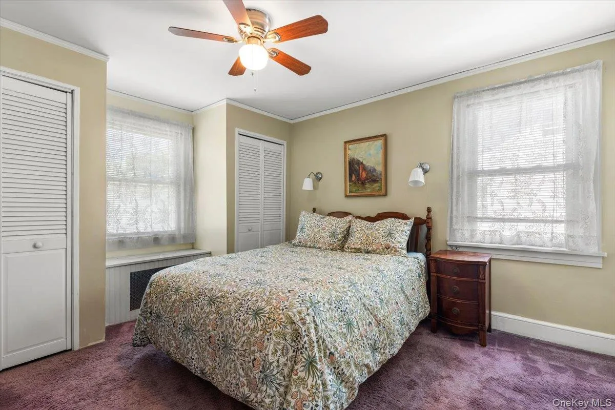 Bedroom featuring two windows with white blinds, purple carpeting, two bi-fold louvered closet doors, a ceiling fan with light fixture, and two wall-mounted sconces Bedroom featuring two windows with white blinds, purple carpeting, two bi-fold louvered closet doors, a ceiling fan with light fixture, and two wall-mounted sconces