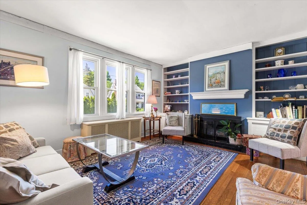 Living room featuring hardwood flooring, a fireplace with a white mantel, and built-in shelving flanking a central wall Living room featuring hardwood flooring, a fireplace with a white mantel, and built-in shelving flanking a central wall