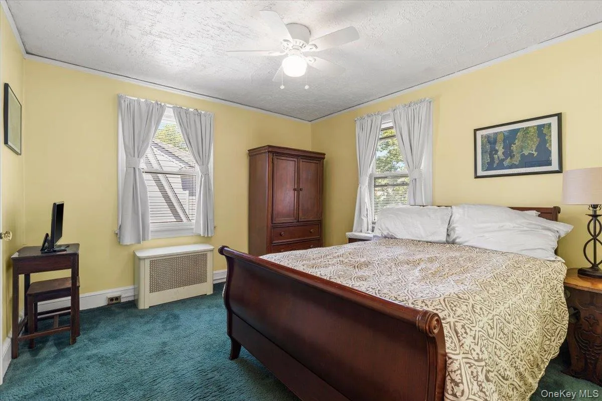 Bedroom featuring two windows with white curtains, teal carpeting, a white ceiling fan, and a radiator beneath one window Bedroom featuring two windows with white curtains, teal carpeting, a white ceiling fan, and a radiator beneath one window