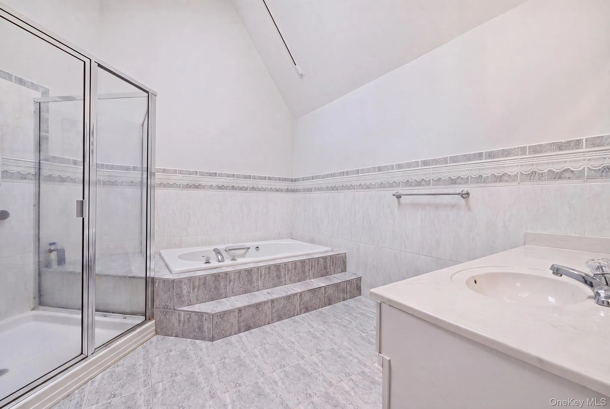 Spacious bathroom featuring a glass-enclosed shower, a stepped platform tub, a single vanity with an integrated sink, tiled walls, and tiled flooring Spacious bathroom featuring a glass-enclosed shower, a stepped platform tub, a single vanity with an integrated sink, tiled walls, and tiled flooring