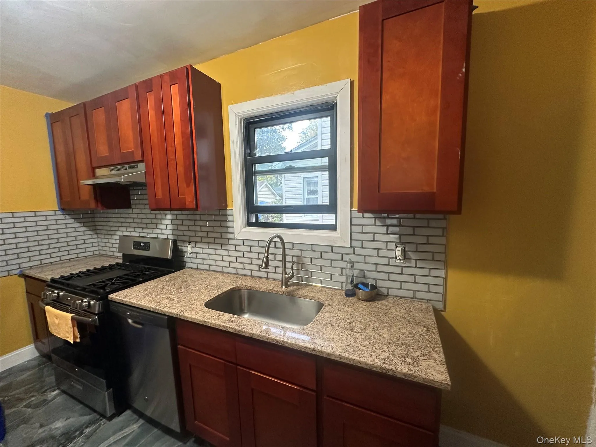 Kitchen with appliances with stainless steel finishes, backsplash, light stone counters, under cabinet range hood, and reddish brown cabinets Kitchen with appliances with stainless steel finishes, backsplash, light stone counters, under cabinet range hood, and reddish brown cabinets