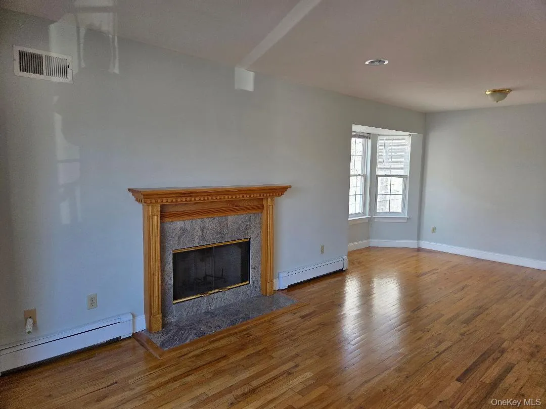 Unfurnished living room featuring wood finished floors, a baseboard radiator, a high end fireplace, and visible vents Unfurnished living room featuring wood finished floors, a baseboard radiator, a high end fireplace, and visible vents