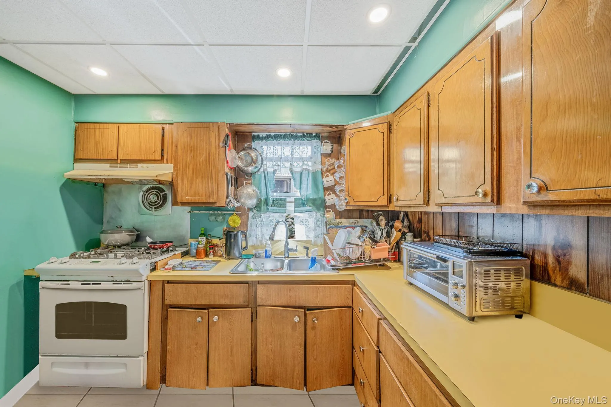 Kitchen with gas range gas stove, light countertops, a paneled ceiling, brown cabinets, and light tile patterned floors Kitchen with gas range gas stove, light countertops, a paneled ceiling, brown cabinets, and light tile patterned floors